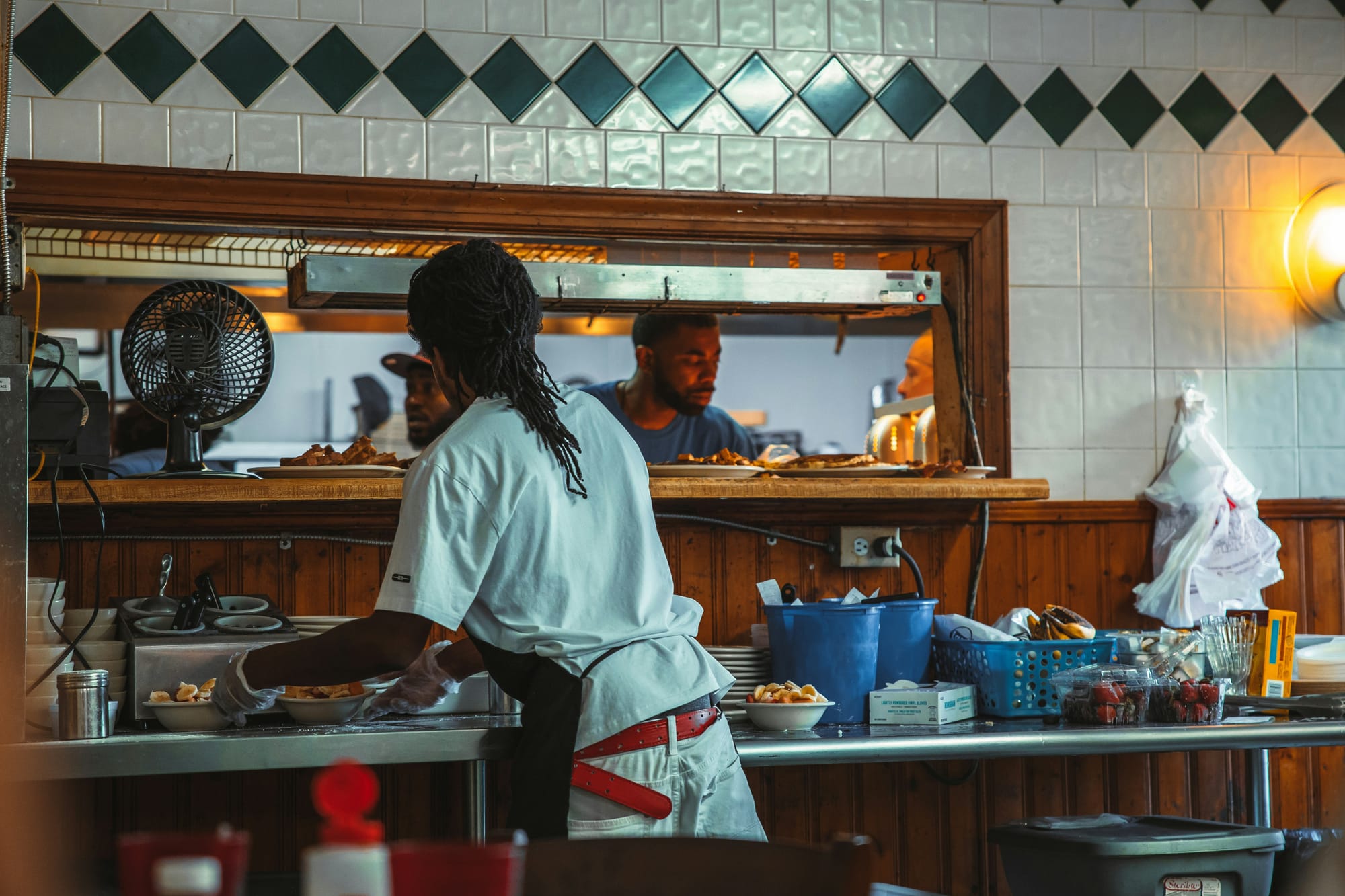A cook plating food in a diner kitchen, two more at the pass window behind